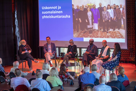 Kuusi ihmistä istuu lavalla panelikeskeskustelussa, taustalla on kuva, jossa lukee uskonnot ja suomalainen yhteiskuntarauha.