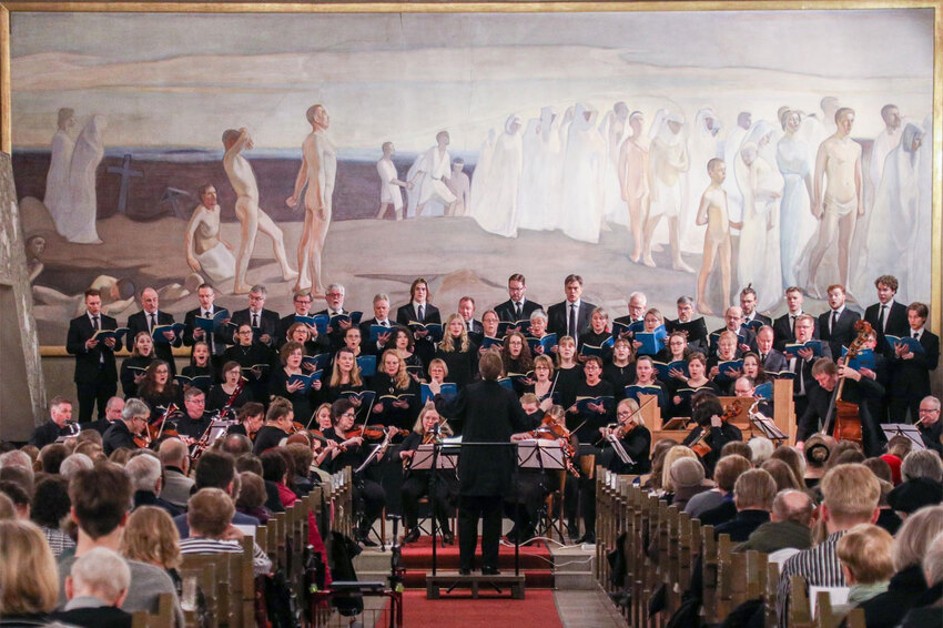 Noin kolmenkymmenen hengen sekakuoro seisoo ihmisä täynnä olevan kirkon alttarioen edessa ja heidän edessään soittaa sinfoniaorkesteri.