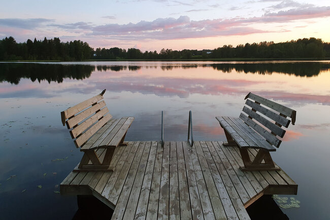 Auringon lasku heijastuu pilvistä järven pintaa violettin sävyin, ja järvessä on puinen laituri.