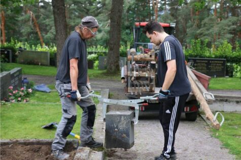 Kaksi miestä kantaa haudan reunuskiveä.