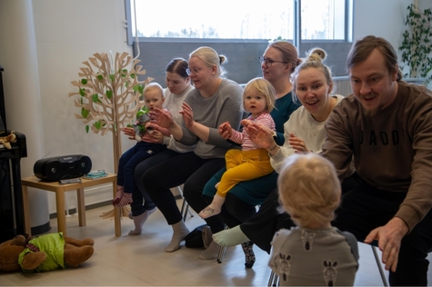 Ryhmä aikuisia ja lapsia istumassa sisätiloissa, osallistuen yhteiseen lauluhetkeen. Aikuiset ja lapset taputtavat käsiään.