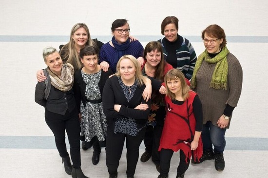 Yläviistosta otettu kuva yhdeksästä naisesta, jotka seisovat. Etummaisena ryhmässä olevalla kädet ovat puuskassa.