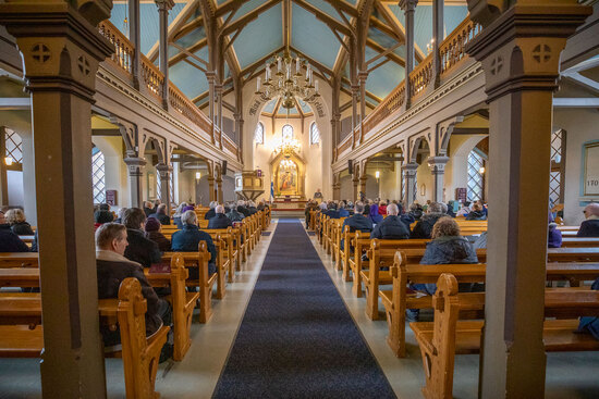 Kauniisti valaistun kirkon etuosassa on Suomen lippu ja pappi on saarnastuolissa, kirkkosalissa istuu ihmisiä harvakseltaan.