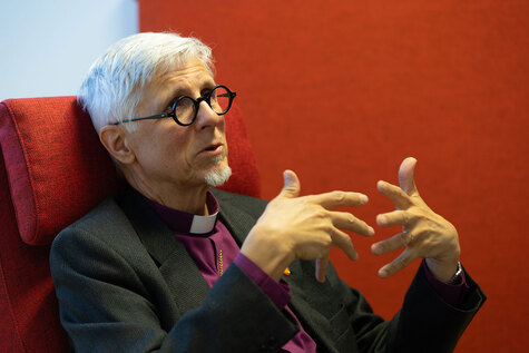 Mies, jolla on piistan puku istuu punaisessa nojatuolissa ja puhuu  myös käsillään.