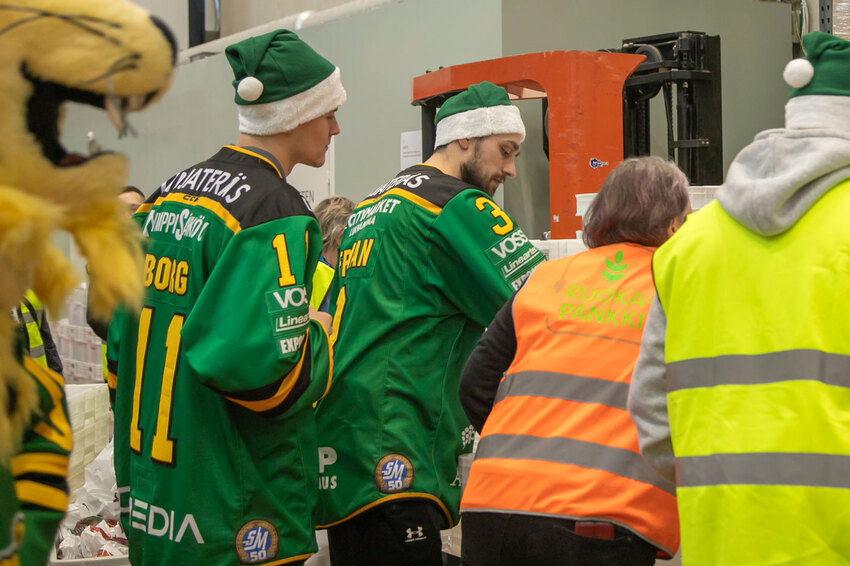 Kaksi Ilves-vaatteisin pukeutunutta miestä nostelee ruokalaatikoita Ruokapankin vaatteisiin pukeutuneen henkilön rinnalla.