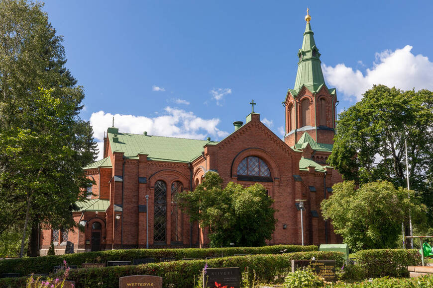 Yleiskuva Messukylän kirkosta, joka on uusgoottista tyyliä edestava punatiilinen kirkko ja kirkko on  vihreiden lehtipuiden ympäröimänä ja etualalla hautausmaan hautakiviä.
