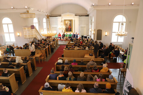 Täyden kirkkosalin etuosassa istuu puolikaaressa seitsemän ihmistä kasvot yleisöön päin.