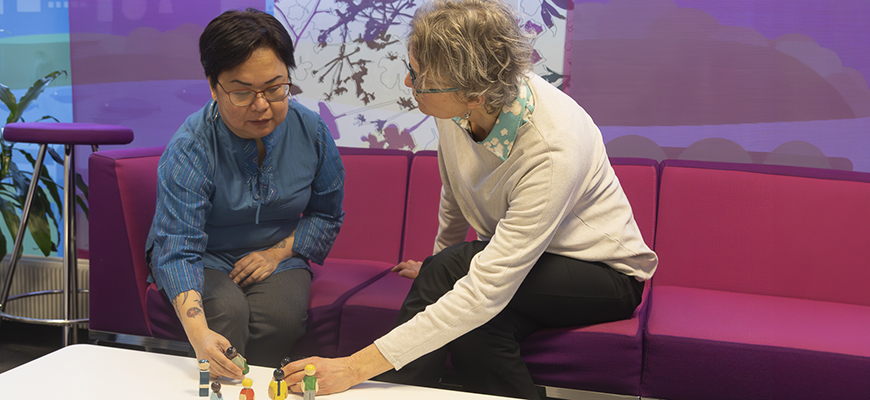 Kaksi ihmistä istuu sohvalla vierekkäin. Edessä pöytä, jossa pieniä puunukkeja, joita ihmiset tutkivat.