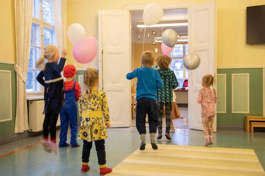 Joukko lapsia juoksee kohti salin avointa ovea ja hyppii ilmaan ilmpallot käsissään.