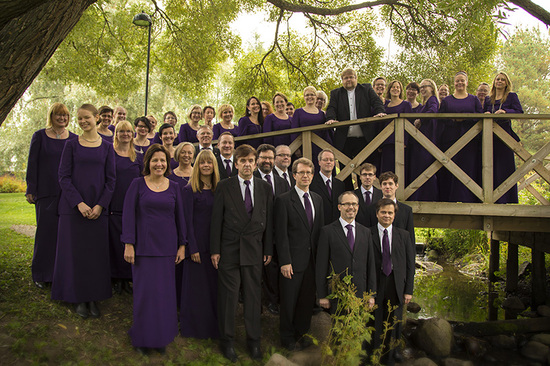 Joukko ihmisiä luonnon keskellä ison puun alla. Osalla on pitkät mekot ja osalla tummat puvut.