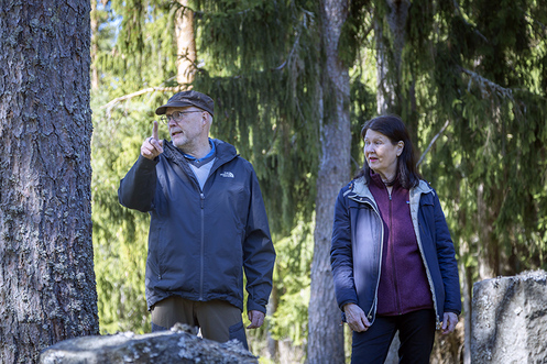 Kaksi ihmistä viittilöi keväällä kuusien katveessa. Etualalla on hautakivien yläreunoja.