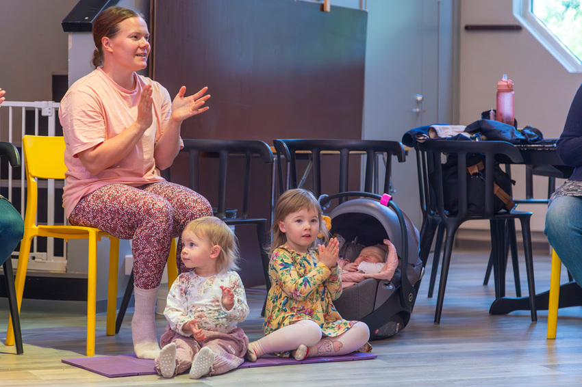 Kaksi pientä lasta istuu kirkkomuskarin lattialla ja taputtaa käsiään yhteen. Vauva nukkuu oikealla puolelle ja äiti istuu vasemmalla puolen penkillä taputtaen käsiään yhteen.