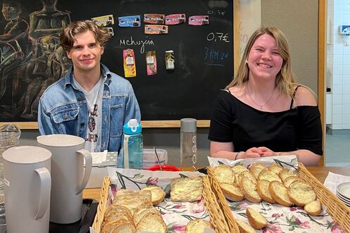 Kaksi nuorta aikuista istuu kahvion tiskin takana. Molemmat hymyilevät.