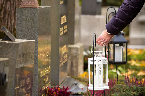 Hautausmaalla henkilö asettaa valkoisen lyhdyn hautakiven viereen, ympärillä syksyisiä kukkia ja muita lyhtyjä.