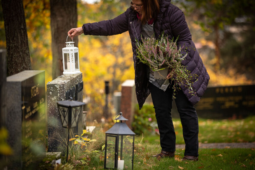 Nainen on laittamassa haudalle kynttilälyhtyä ruskan värittämällä hautausmaalla.