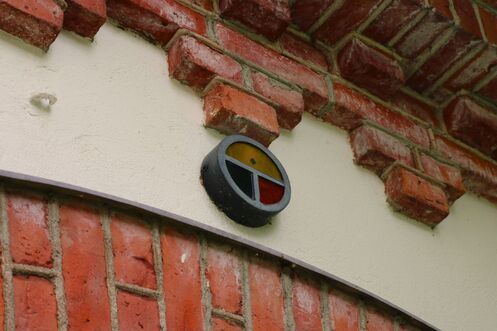 Tiiliseinän keskellä kulkee vaakasuuntainen valkoinen kaistale. Valkoisen kaistaleen yläpuolella on punatiilinen holvikaari. Holvikaaren keskellä on pyöreä merkki, joka on jaettu kolmeen osaan: yläosa on keltainen, vasen alaosa sininen ja oikea alaosa punainen. Osat ovat värjättyä lasia.