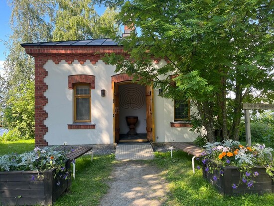 Pieni tiilinen kappeli kutsuvasti ovi avoinna, keskellä kesäistä vehreyttä, etualalla värikkäitä kukkia.