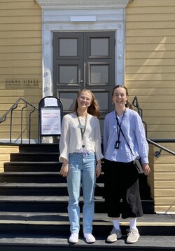 Taustalla näkyy vanha keltainen puukirkko, sen portailla hymyilee kaksi nuorta kirkon kesäopasta auringonpaisteessa