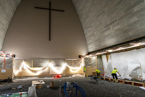 Molemmilta reunoiltaan ylöspäin kapenevassa ja kaareutuvassa takaseinässä on iso risti. Lattian maapohja on paljastunut. Keskellä on rakennustyömaan välineistöä ja seinän vieressä lapioi kaksi henkilöä.