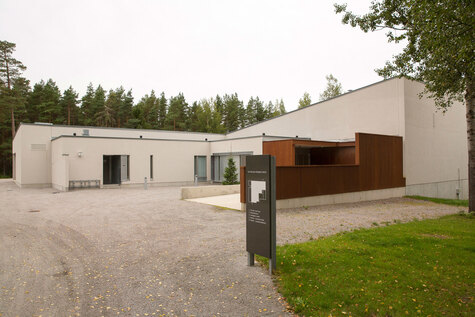 Vaaleanharmaa moderni rakennus metsäisesä ympäristössä, jossa on monimutkainen muotoilu ja suuria ikkunoita.