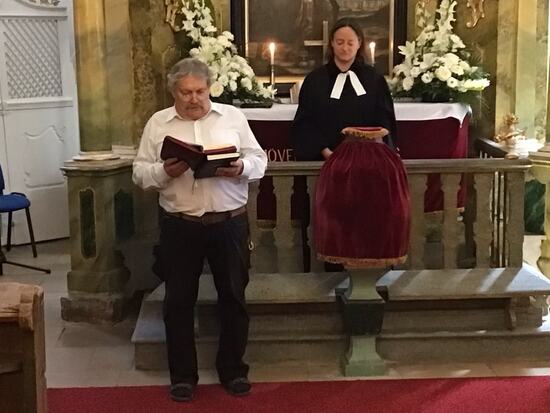 Kaksi henkilöä seisoo kirkon alttarin edessä hartaan tunnelman vallitessa. Etualalla oleva henkilö lukee Raamattua keskittyneesti. Taustalla toinen henkilö seisoo alttarin takana, jonka päällä on punainen kangas, kukka-asetelmia ja palavia kynttilöitä. Alttarin taustaa koristavat maalaukset ja koristeelliset yksityiskohdat, jotka luovat arvokkaan ja hengellisen ilmapiirin.