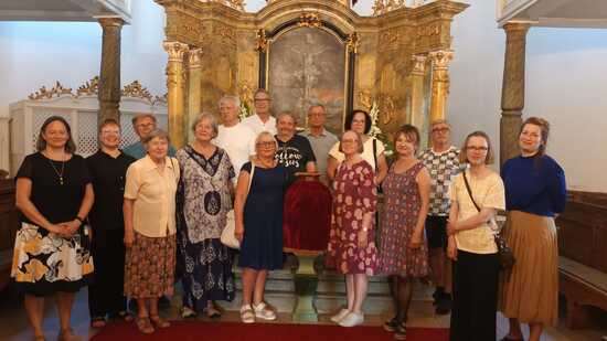 Ryhmä miehiä ja naisia seisoo kirkon alttarilla, pukeutuneina kesäisiin vaatteisiin. Taustalla kohoaa koristeellinen alttaritaulu, jota reunustavat kultaiset yksityiskohdat. Keskellä oleva henkilö pitelee punaista samettikangasta, joka tuo juhlavuutta tilanteeseen. Tunnelma viittaa yhteiseen hetkeen kirkossa osana yhteistä matkaa.