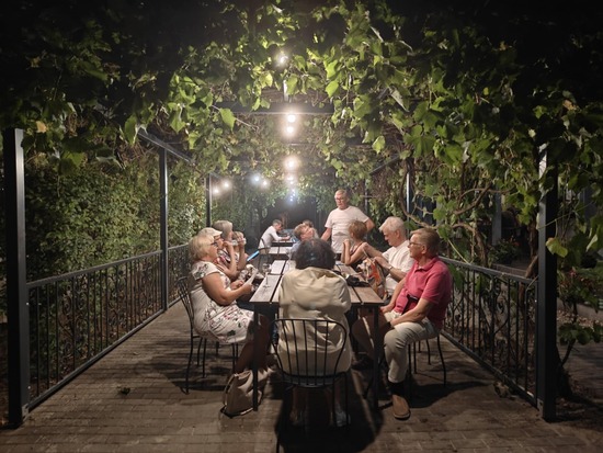 Ryhmä ihmisiä istuu pitkän puupöydän äärellä vehreän pergolan alla, viiniköynnöksen suojassa. Pimenevä ilta  ja ympäröivä vihreys luovat rennon ja kutsuvan tunnelman, jota täydentävät katosta roikkuvat valot. Keskustelut soljuvat ja yhdessäolo on lämminhenkistä.