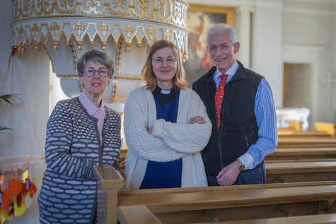 Kaksi naista ja yksi mies seisoo kirkossa, penkkien välissä ja hymyilevät kameralle.