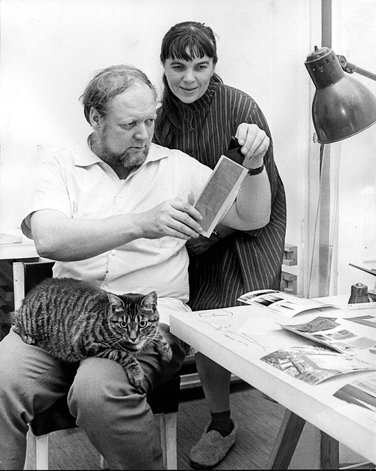 Raili ja Reima Pietilä tutkivat valokuvia pöydän ääressä. Raidallinen kissa istuu Reima Pietilän sylissä.
