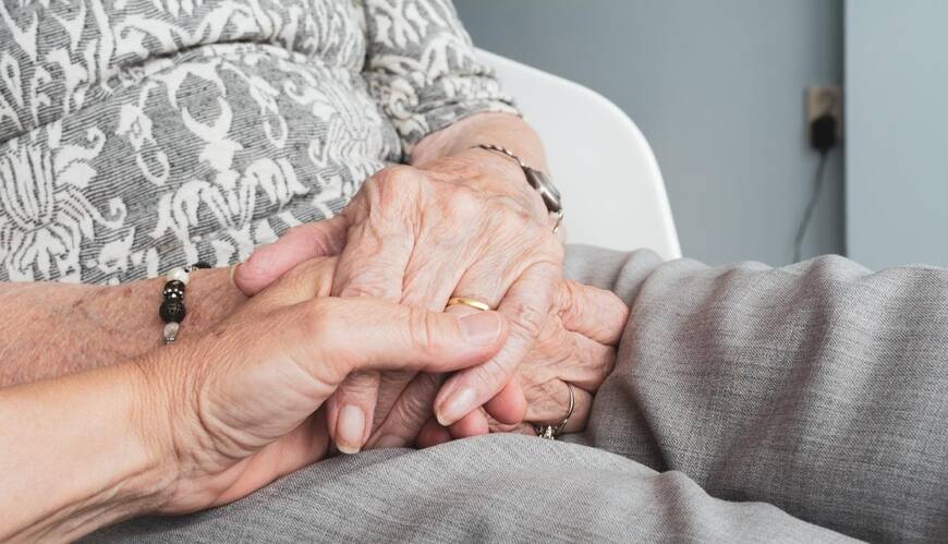 Etualalla näkyy nuoremman ihmisen kädet, jotka ovat lempeästi vanhemman, ryppyisten käsien päällä. Vanhemmalla henkilöllä on harmaa-huopakuvioinen paita ja harmaat housut. Taustalla näkyy vaaleansininen seinä. Tunnelma on lämmin, lohduttava ja myötätuntoinen.