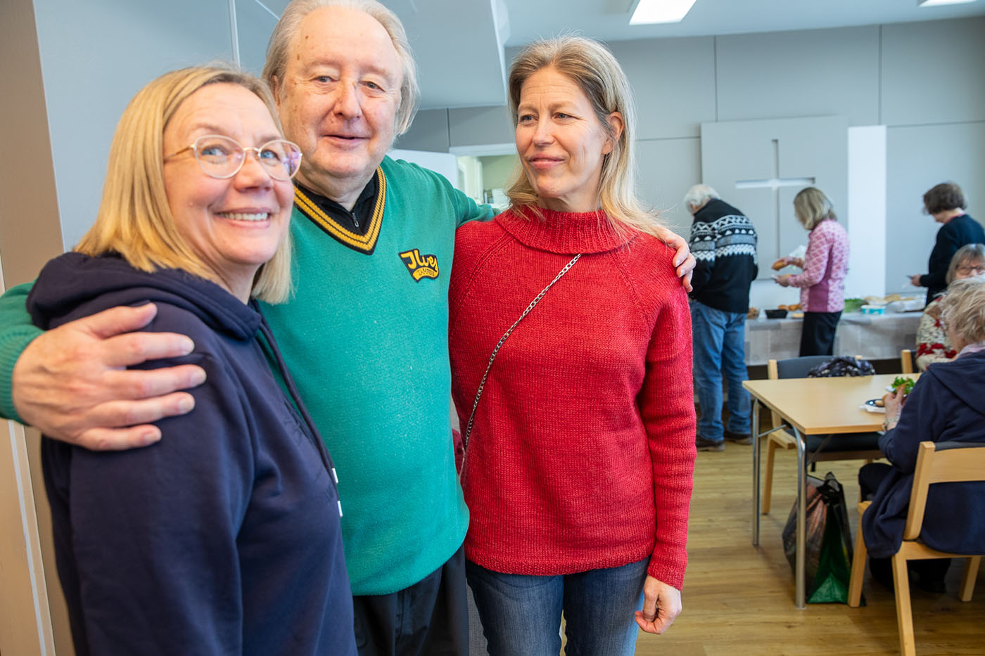 Kaksi naista miehen molemmin puolin. Mies on kietonut kätensä naisten ympärille. Kaikki hymyilevät. Taustalla muita ihmisiä.
