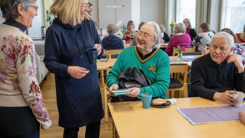 Mies istuu pöydän ääressä, edessään kahvikuppi ja syötävää. Mies pitää kädessään kahta kirjaa ja esittelee niitä edessään seisovalle naiselle. Molemmat hymyilevät. Taustalla eri ikäisiä ihmisiä seurakuntasalissa istumassa ja seisomassa pöytien ääressä.