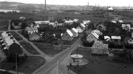 Mustavalkoinen kuva 1930-luvulta. Puutaloja ja hiekkateitä. Taustalla tehtaiden piippuja ja kaupunki.