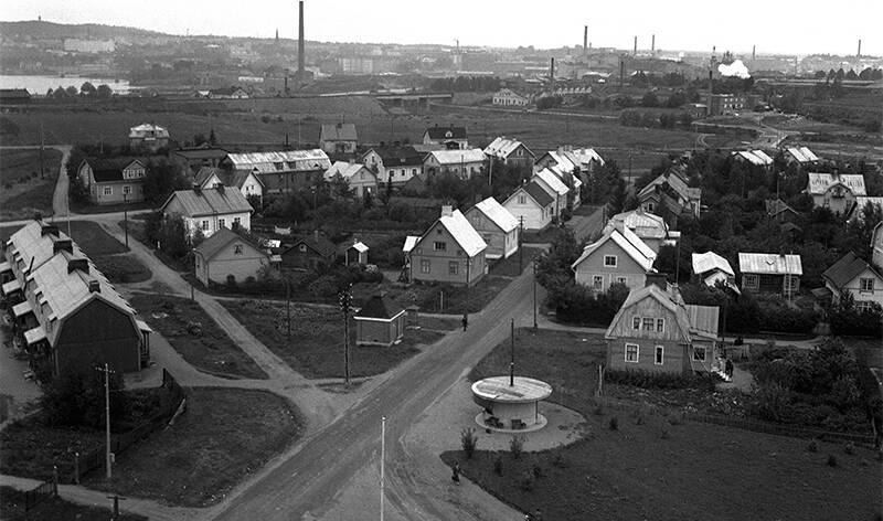 Mustavalkoinen kuva 1930-luvulta. Puutaloja ja hiekkateitä. Taustalla tehtaiden piippuja ja kaupunki.
