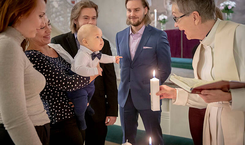 Aamukaste-tilaisuus. Pappi ilmeilee sylivauvalle.