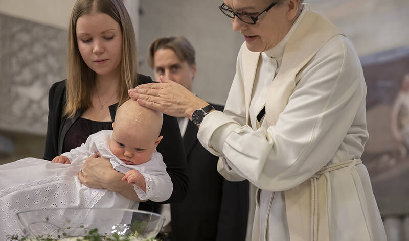 Nuori nainen pitää sylissään vauvaa, jonka pään päälle pappi on laskenut kätensä. Taustalla on kolmas henkilö.