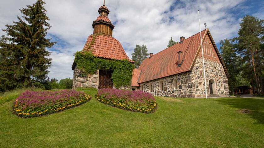 The old grey stone church, with a red tiled roof, sits atop a hill, and in front of the church, there is a green grassy area with flower beds.