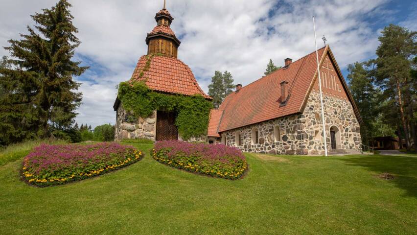 The old grey stone church, with a red tiled roof, sits atop a hill, and in front of the church, there is a green grassy area with flower beds.