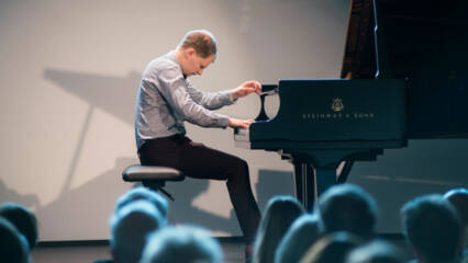 Sivuprofiili pianoa soittavasta henkilöstä. Kuvan etualalla on selin kameraan olevaa yleisöä.