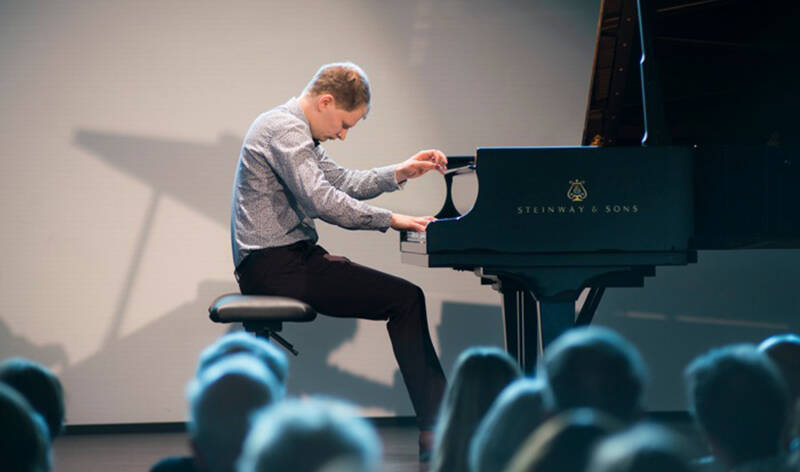 Sivuprofiili pianoa soittavasta henkilöstä. Kuvan etualalla on selin kameraan olevaa yleisöä.