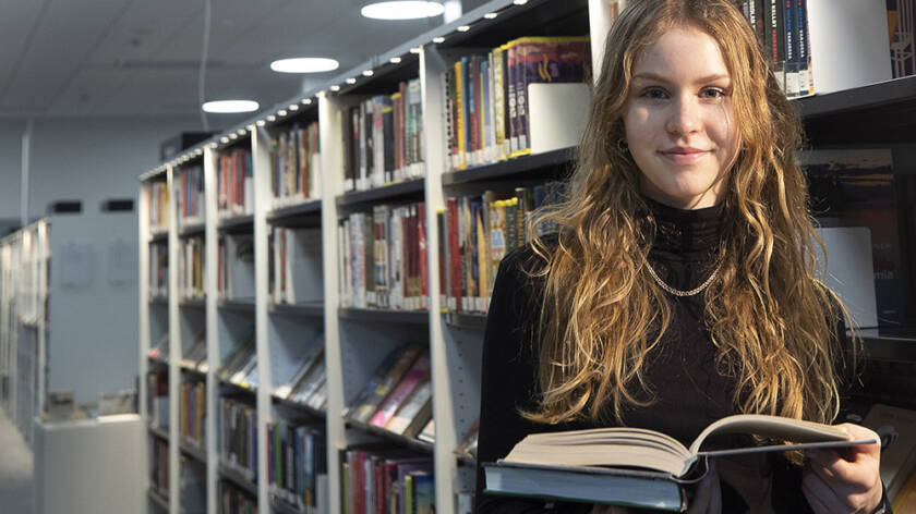 Puolivartalokuva teinitytöstä, joka seisoo kirja käsissään kirjastossa.
