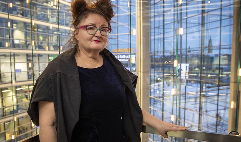 Silmälasipäinen nainen hymyilee ja seisoo ison, avaran rakennuksen ylemmässä kerroksessa ja pitää toista kättä kaiteen päällä. Takana näkyy valtavamäärä rakennuksen ikkunoita.