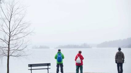 Kolme ulkoiluvaatteisiin pukeutunutta ihmistä seisovat jäätyneen järven rannalla ja katsovat lumiselle ulapalle.