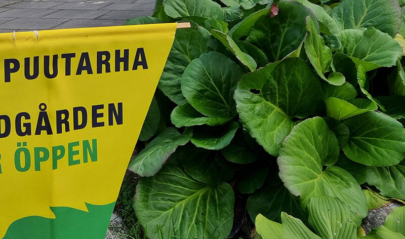 Kyltti, josta näkyvät tekstit puutarha, gården ja öppen. Vieressä on vehreää kasvustoa.