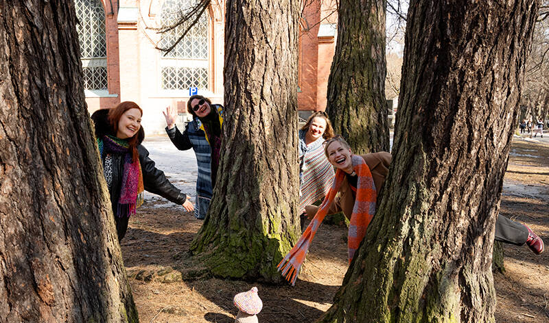 Puitten takaa kurkistaa neljä iloista naista ja heidän keskellä maassa on neulottu sieni.