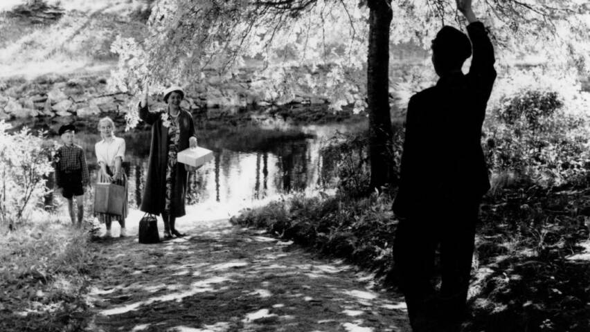 Mustavalkoinen kuva Matti Kassilan elokuvasta Elokuu (1956). Päähenkilö tervehtii perhettään veden rannalla.