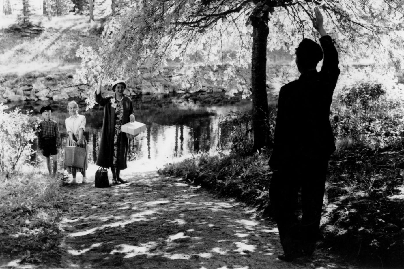 Mustavalkoinen kuva Matti Kassilan elokuvasta Elokuu (1956). Päähenkilö tervehtii perhettään veden rannalla.