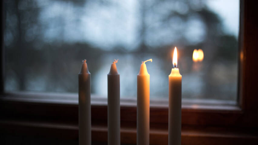 Neljä kynttilää rivissä ikkunan edessä, takana pimenevä metsä, oikean puoleissa kynttiläss pala liekki.