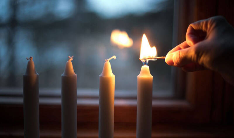 Neljä kynttilää rivissä ikkunan edessä, takana pimenevä metsä, oikean puoleista kynttilää ollaan sytytämässä kädessä oleva tulitikulla.