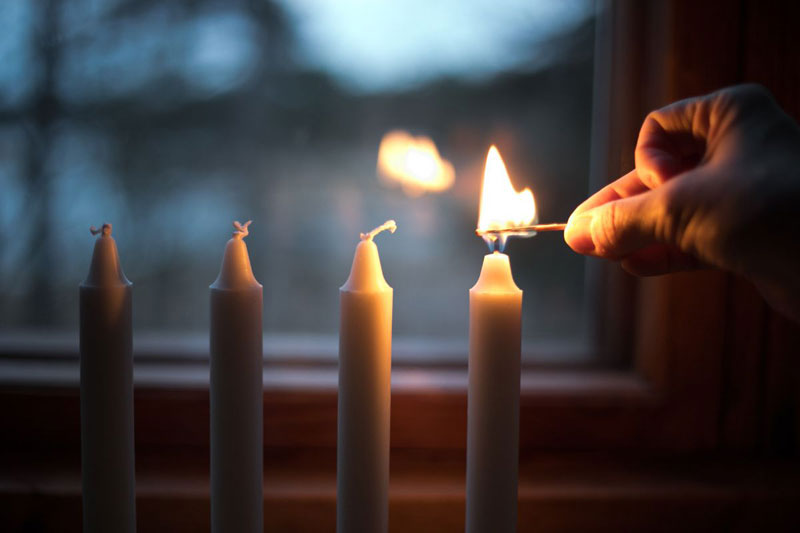 Neljä kynttilää rivissä ikkunan edessä, takana pimenevä metsä, oikean puoleista kynttilää ollaan sytytämässä kädessä oleva tulitikulla.